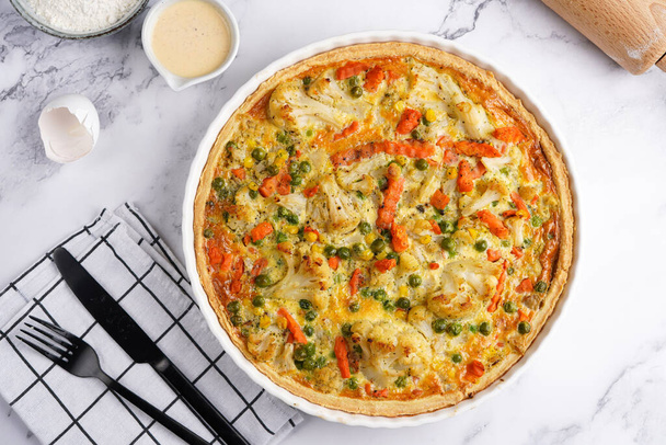 Baked french vegetable pie quiche filled with carrots, peas and cheese on a marble surface, cracked egg shell, flour, cream, top view - Foto, Imagem