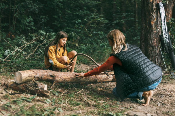 Een moeder en haar dochter zagen een omgevallen boom voor een vuur. Brandhout oogsten, kampvuur, picknick - Foto, afbeelding