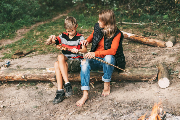 Een moeder en haar tienerzoon zitten op een boom in campings en proberen gegrilde worsten gekookt op een vuur - Foto, afbeelding