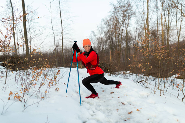 冬に森の中を走る前に女性ランナーが暖まる。寒い冬の日にトレイルランニングの前にストレッチトレッキングポールを持つスポーツ女性. - 写真・画像