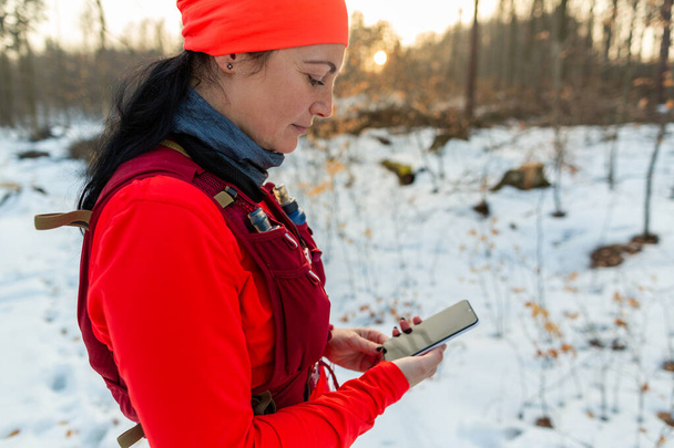 Mujer revisando el móvil mientras corre en el bosque. Corredor femenino sosteniendo el teléfono celular en las manos en el frío día de invierno. - Foto, Imagen