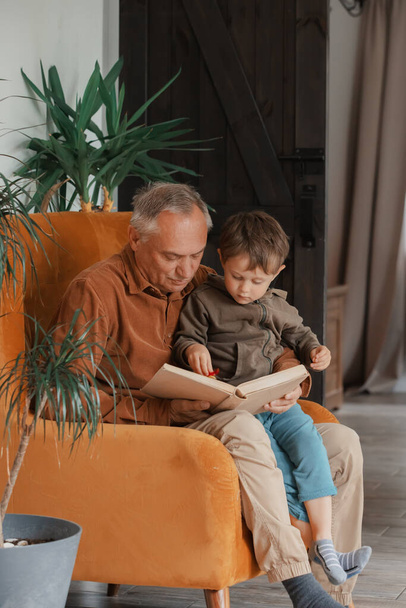 Ο παππούς διάβαζε ένα βιβλίο με τον εγγονό του στο σπίτι - Φωτογραφία, εικόνα