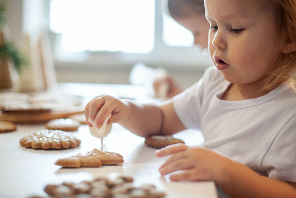 children decorate Christmas gingerbread at home. A boy and a girl paint with cornets with sugar icing on cookies. New Years decor, branches of a Christmas tree. - Фото, изображение