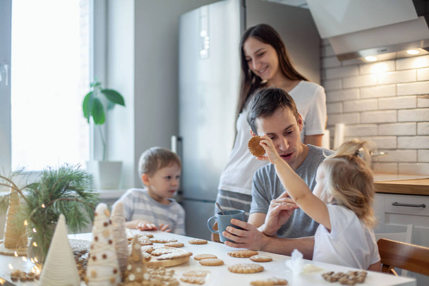 Dad, mom and children decorate Christmas gingerbread at home. A boy and a girl paint with cornets with sugar icing on cookies. New Years decor, branches of a Christmas tree. - Zdjęcie, obraz
