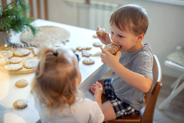 Boy and girl decorate and eating Christmas gingerbread at home. A boy and a girl paint with cornets with sugar icing on cookies. New Years decor, branches of a Christmas tree. - Foto, immagini