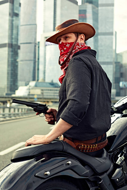 Portrait of stylish male dressed in a black leather jacket and bandana with gun sitting on his custom-made retro motorcycle, looking away, buildings of the big city on background. - Foto, Bild