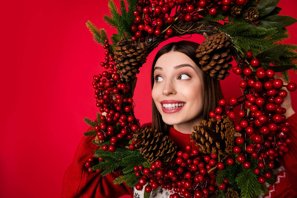 Photo of adorable cute young lady wear print sweater smiling looking inside christmas wreath empty space isolated red color background - Foto, afbeelding