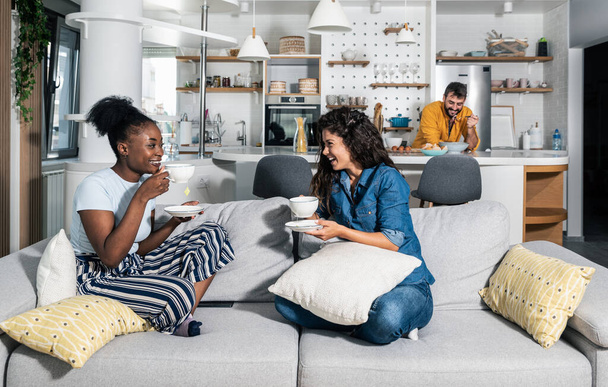 White woman tells her black friend that her boyfriend is a good lover while they sit on the sofa and drink tea or coffee and man stands in the kitchen and eating without knowing what they are talkin - Fotoğraf, Görsel