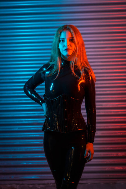 Red-haired girl in latex, a black top and tight pants, on a metallic background illuminated with red and blue LEDs - Photo, image