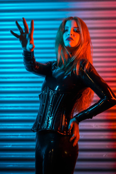 Red-haired girl wearing latex, a black top and tight pants, on a metallic background illuminated with red and blue LEDs, making the stop gesture - Valokuva, kuva