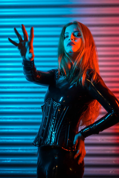 Red-haired girl wearing latex, a black top and tight pants, on a metallic background illuminated with red and blue LEDs, making the stop gesture - Fotó, kép