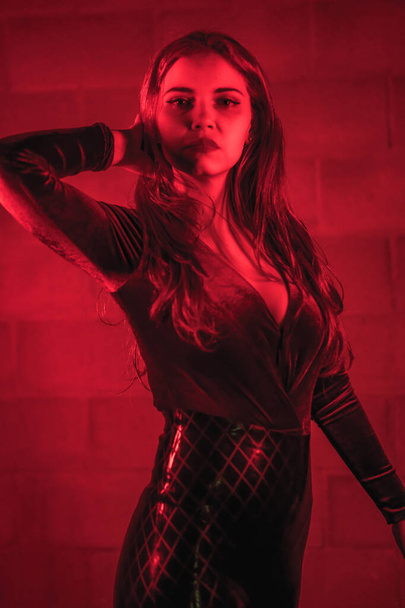 Red-haired girl in latex, brown and black tight suit on a brick wall, posed at night illuminated with red light - Foto, afbeelding