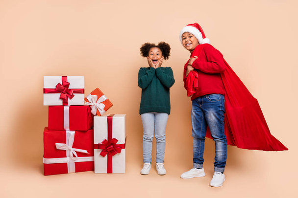 Photo of two kids celebrate new year boy carry sack gifts wear x-mas hat jumper isolated beige color background - 写真・画像