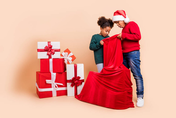 Photo of two curious kids look presents bag shocked face wear x-mas hat jumper isolated beige color background - Фото, изображение
