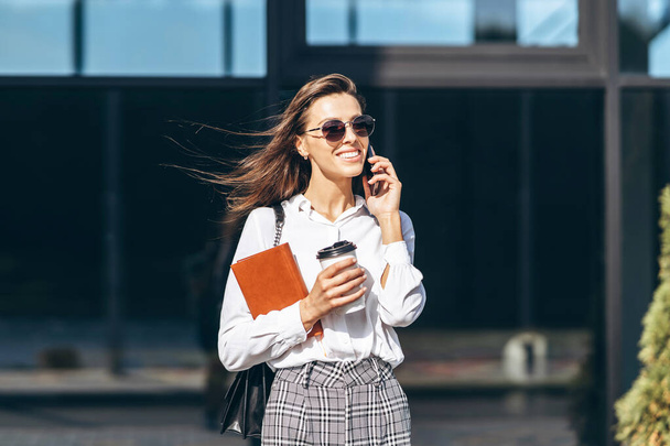 Business woman walking and talking on the cellphone outdoors near modern business center with notebook - Foto, Imagen
