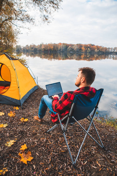 mies istuu tuolissa lähellä syksyn järvi työskentelee kannettavan freelance camping käsite - Valokuva, kuva