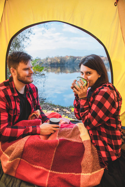 couple sitting in the tent drinking warm tea looking at autumn lake. camping concept - Photo, Image