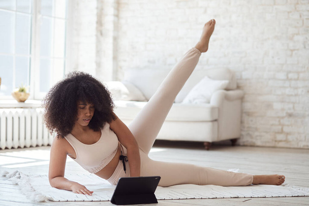 Αθλητική νεαρή μαύρη γυναίκα βλέποντας βίντεο φροντιστήριο στο tablet κάνει ασκήσεις στο σπίτι με μεγάλα παράθυρα το πρωί. Κοίτα. - Φωτογραφία, εικόνα