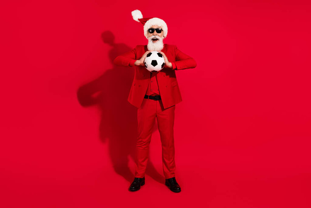 Full size photo of cool grey hair aged man hold ball wear santa cap red suit eyewear isolated on red color background - 写真・画像