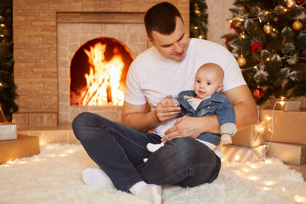 Young adult brunet father wearing jeans and white casual t shirt holding small daughter in hands, posing at home near fireplace and Christmas tree, spending winter holidays with family. - Foto, Imagem