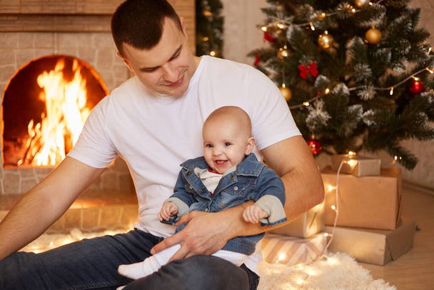 Handsome brunet father holding his small infant daughter in hands, looking at his cute charming kid, sitting near fireplace and xmas tree at home, happy new year. - Foto, imagen