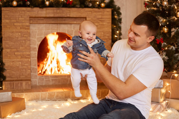 Indoor shot of handsome father wearing white casual t shirt holding little toddler daughter in hands while sitting near fireplace and xmas tree at home, happy new year. - Fotó, kép
