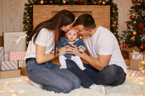 Portrait of young family kissing little daughter, sitting on the floor near Christmas tree and fireplace, spending and celebrating winter holidays together at home. - Photo, image