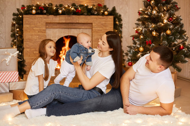 Portrait of happy family of father, mother and two daughters sitting on floor on soft carpet near fireplace and xmas tree, celebrating winter holidays, happy new year. - Φωτογραφία, εικόνα
