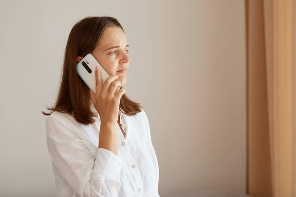 Side view of positive woman wearing white cotton shirt standing near window with beige cottons and talking phone, looking away, having calm facial expression. - Foto, imagen