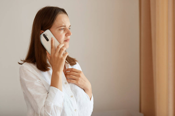 Portrait of upset female with dark hair wearing white cotton shirt standing indoor and talking phone, hearing bed news, having sad facial expression, looking away. - Foto, Imagen