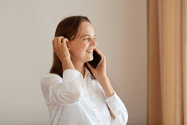 Side view portrait of positive woman standing near window with beige curtains, young adult girl wearing white cotton shirt talking via cell phone, hearing good news. - Photo, Image