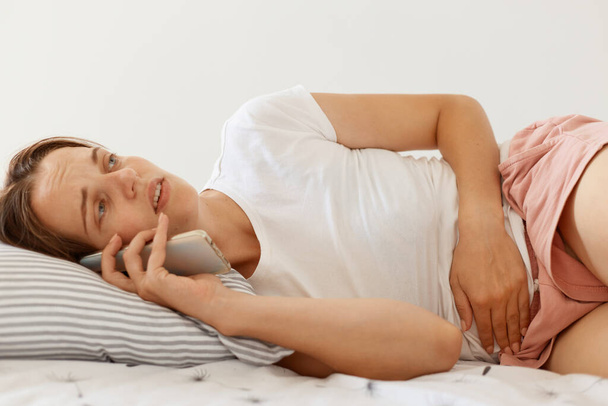 Ill woman with dark hair wearing white casual t shirt lying in bed and talking via smart phone with her doctor, having problems with stomach, suffering from abdominal pain. - Foto, Bild