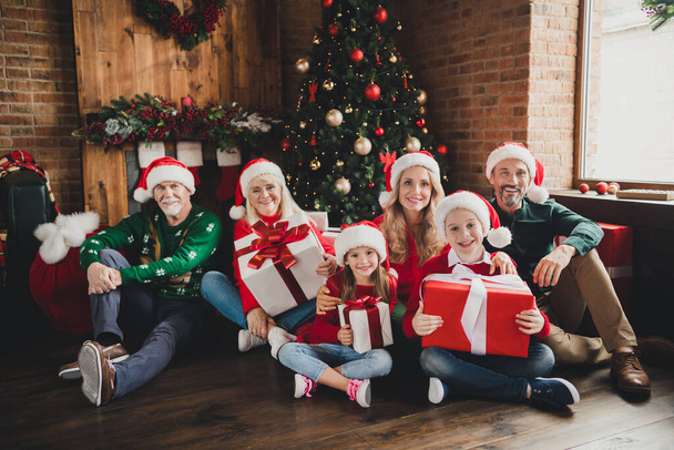 Retrato de familia alegre atractiva con sombreros celebración de regalos de diciembre sentado en el árbol del piso en casa loft casa interior - Foto, Imagen