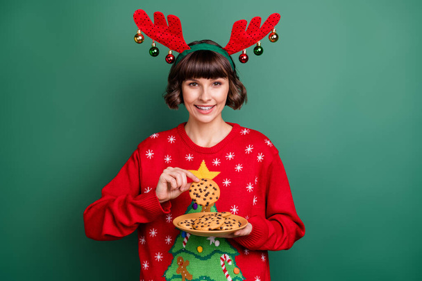 Photo of sweet adorable young woman dressed red pullover antlers smiling holding new year gingerbreads isolated green color background - 写真・画像