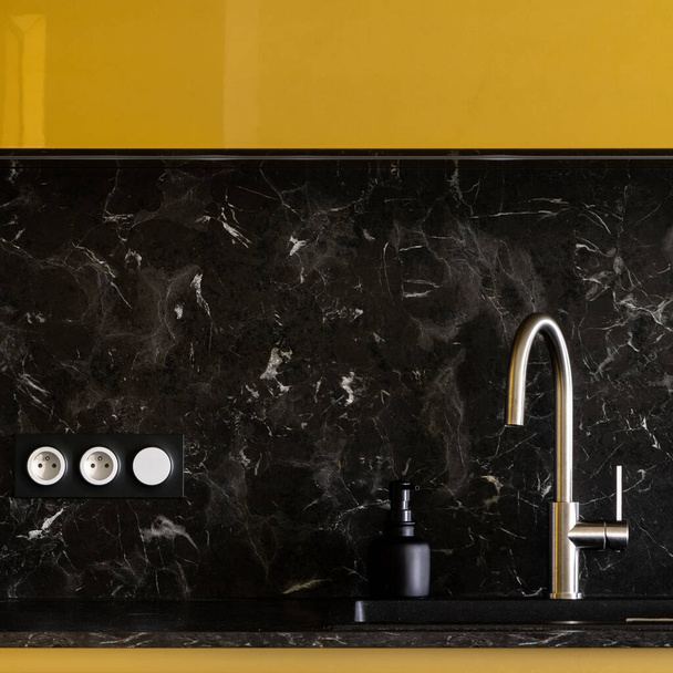 Close-up on silver tap in black sink in elegant kitchen with gold cupboards and black marble style backsplash tile - Photo, image