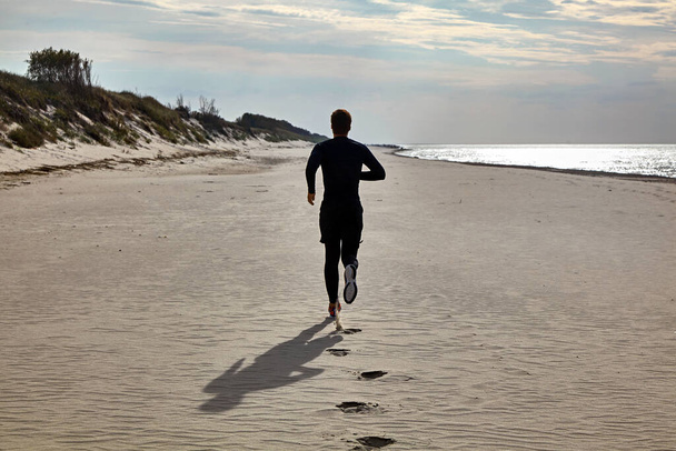 A young man in black sports gear and orange sneakers is jogging on the beach - Foto, Imagem