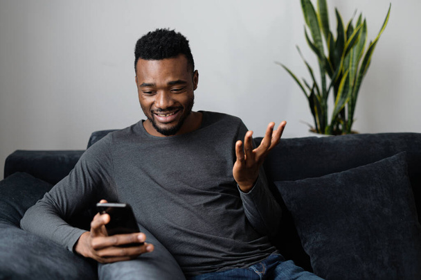 Surprised African-American guy sitting on the couch and holding smartphone - Фото, зображення