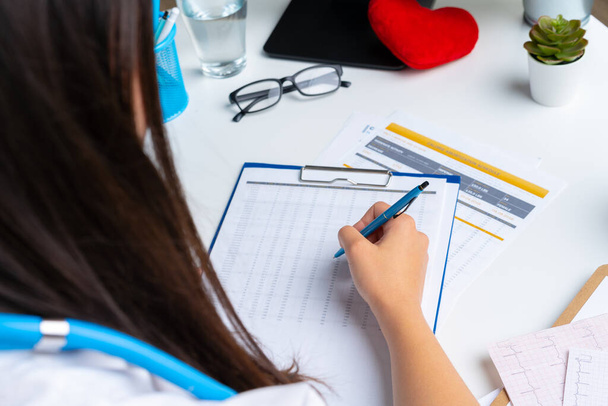 Woman doctor taking notes on clipboard while sitting at her table in office - Foto, Bild