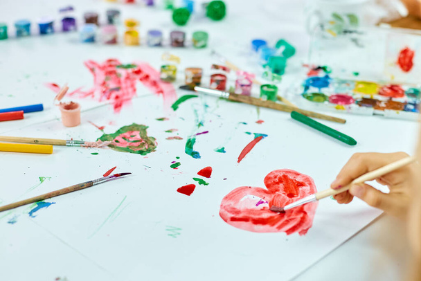 Roztomilé dítě sedí u stolu a kreslí červené srdce na bílém papíru, Umělecká škola koncept, dívka dítě malování s akvarely doma - Fotografie, Obrázek