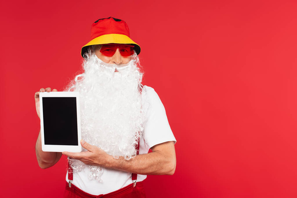 Bearded santa claus in sunglasses holding digital tablet with blank screen isolated on red  - Photo, image