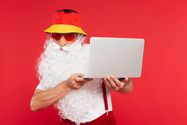 Santa Claude en panama et lunettes de soleil tenant ordinateur portable isolé sur rouge  - Photo, image