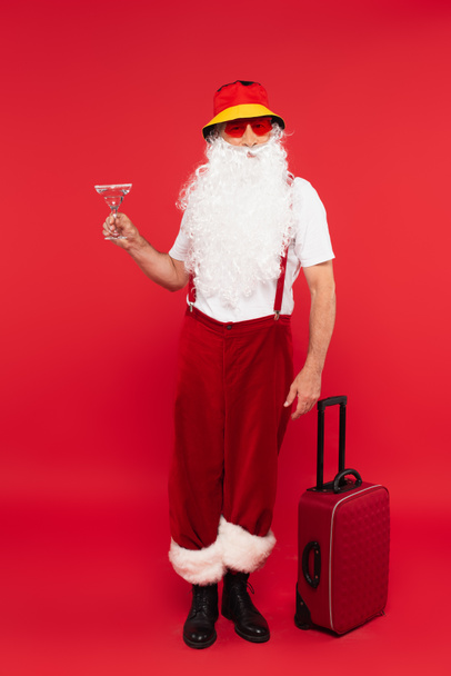 Άγιος Βασίλης σε γυαλιά ηλίου κρατώντας κοκτέιλ κοντά σε βαλίτσα σε κόκκινο φόντο - Φωτογραφία, εικόνα