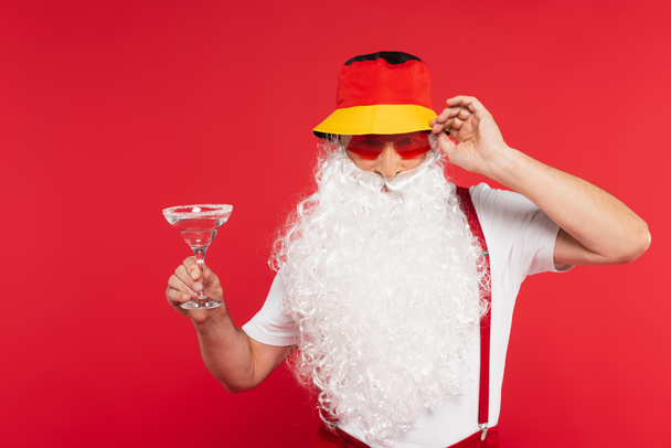 Santa claus in panama and sunglasses holding glass of cocktail isolated on red  - Zdjęcie, obraz