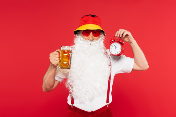 Santa claus in sunglasses holding vintage alarm clock and glass of beer isolated on red  - Φωτογραφία, εικόνα