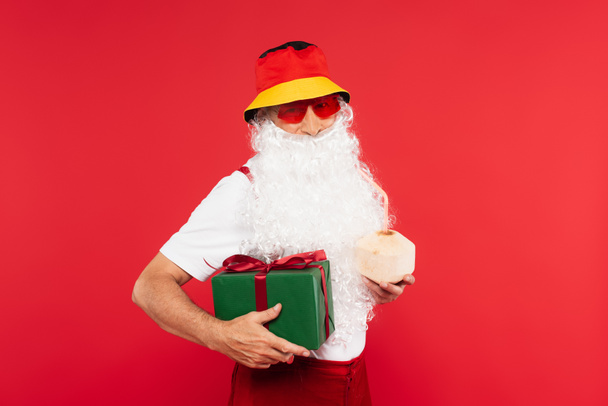 Santa Claude dans des lunettes de soleil tenant boîte cadeau et cocktail en noix de coco isolé sur rouge  - Photo, image