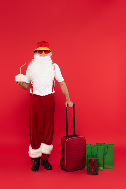 Santa claus in sunglasses holding cocktail in coconut and suitcase near shopping bags and present on red background - Photo, Image
