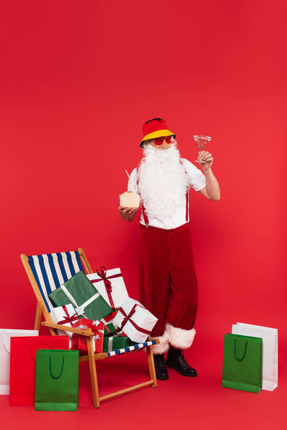 Санта-Клаус держит коктейли рядом с сумками и подарками на шезлонге на красном фоне - Фото, изображение
