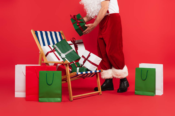 Vista ritagliata di Natale padre tenendo presente vicino sedia a sdraio e borse della spesa su sfondo rosso - Foto, immagini