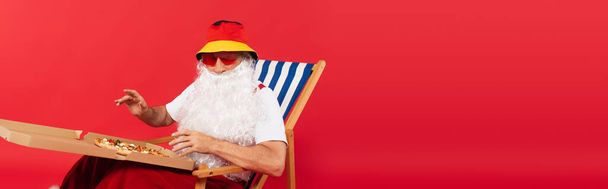 Père Noël dans des lunettes de soleil tenant la boîte à pizza tout en étant assis sur la chaise longue isolé sur rouge, bannière  - Photo, image