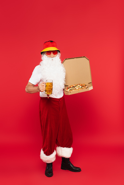 Father christmas in sunglasses holding mug of beer and pizza on red background - Photo, Image